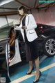 A woman in a white blazer and black dress standing next to a car.